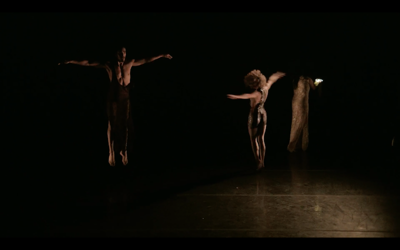 A shirtless man with his arms outstretched in the semi darkness. Another figure with her back to the audience also outstretches her arms